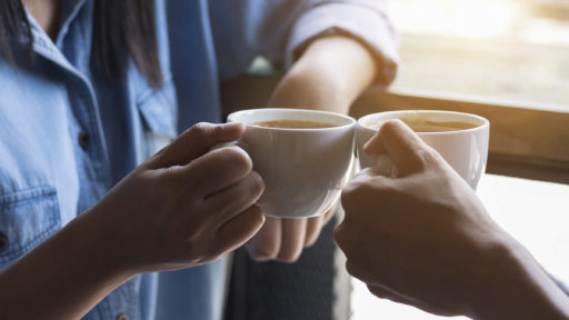 Two women in a coffee shop lifting coffee mugs to cheers.