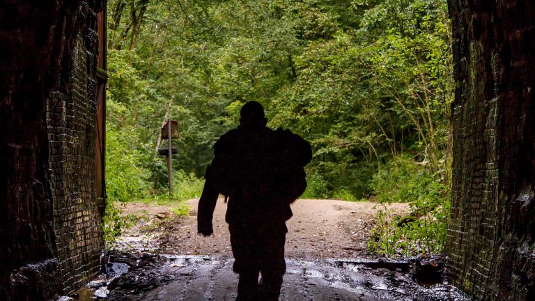 A soldier walks through a dark tunnel with a light and tree filled opening.